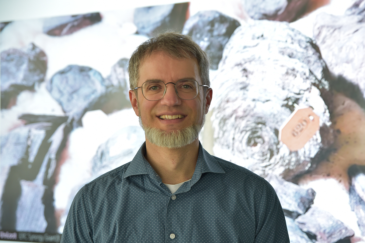 Ein Wissenschaftlermit Bart vor einer Leinwand. Im Hintergrund verkohlte Papyri.