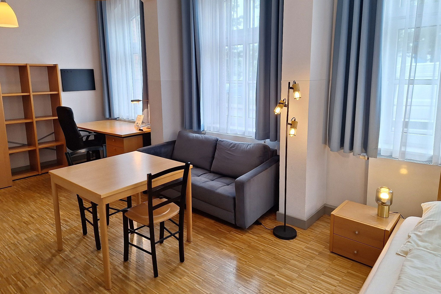View of a room with couch, table, bed, desk, and shelves.