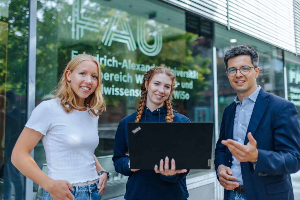 Drei Personen stehen mit Laptop in der Hand vor einer Fensterfront mit FAU-Logo.