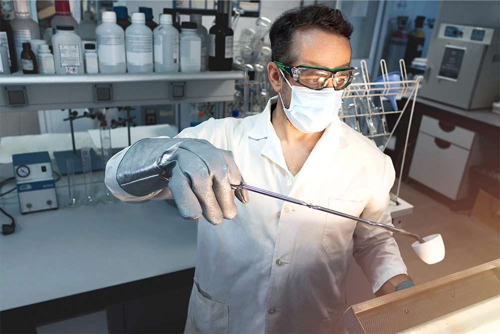 Ein Mann mit Brille und OP-Maske in Labor: Mit Handschuh und einem langen Instrument öffnet er einen Ofen.