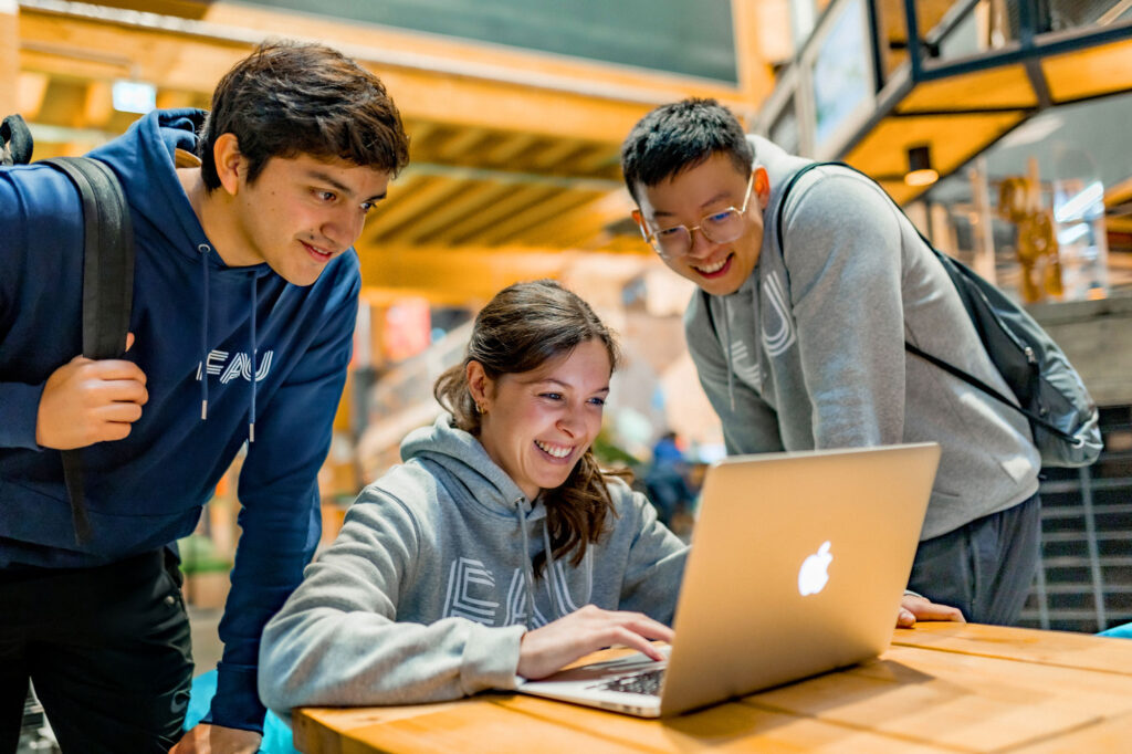 Two students from Friedrich-Alexander-Universität Erlangen-Nürnberg are leaning over a laptop on a desk in front of a third student.