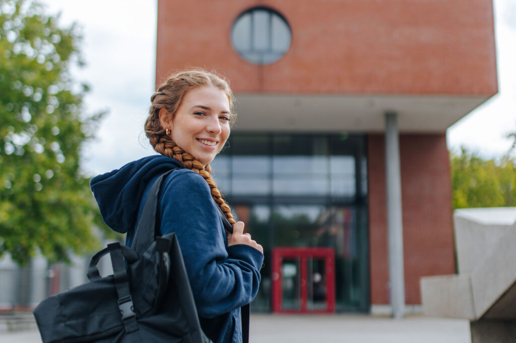 Eine Studentin trägt eine Tasche und steht vor einem Gebäude.