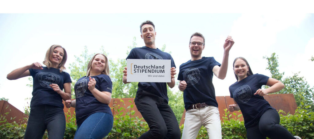 Fünf Studierende springen in die Luft, ein Student hält ein Schild mit der Aufschrift des Deutschland Stipendiums hoch