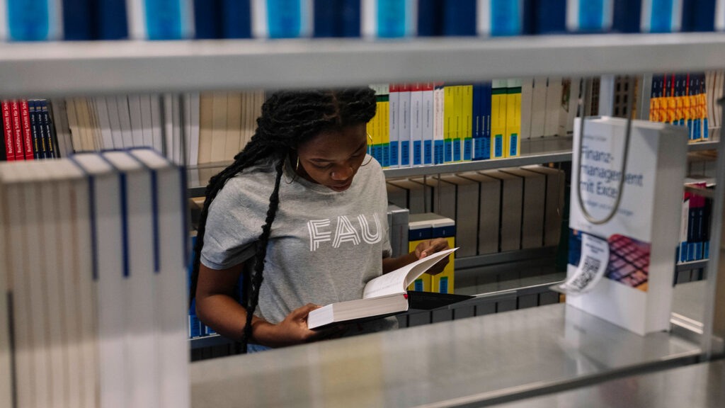 Eine junge Frau steht in einer Bibliothek und liest in einem Buch.