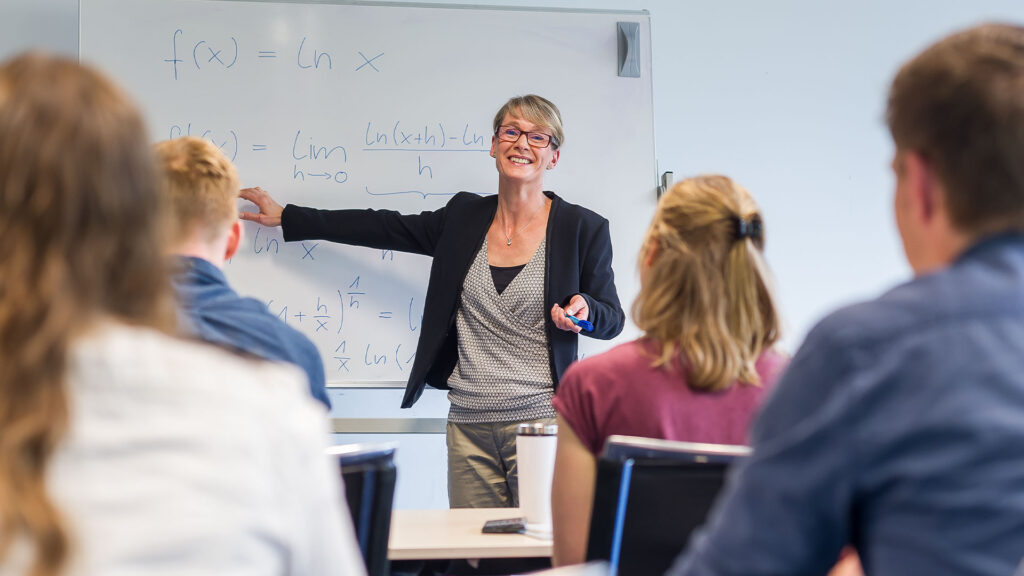 Eine Frau zeigt auf ein Whiteboard, auf das sie etwas geschrieben hat. Sie schaut zu ihren Studenten und erklärt etwas.