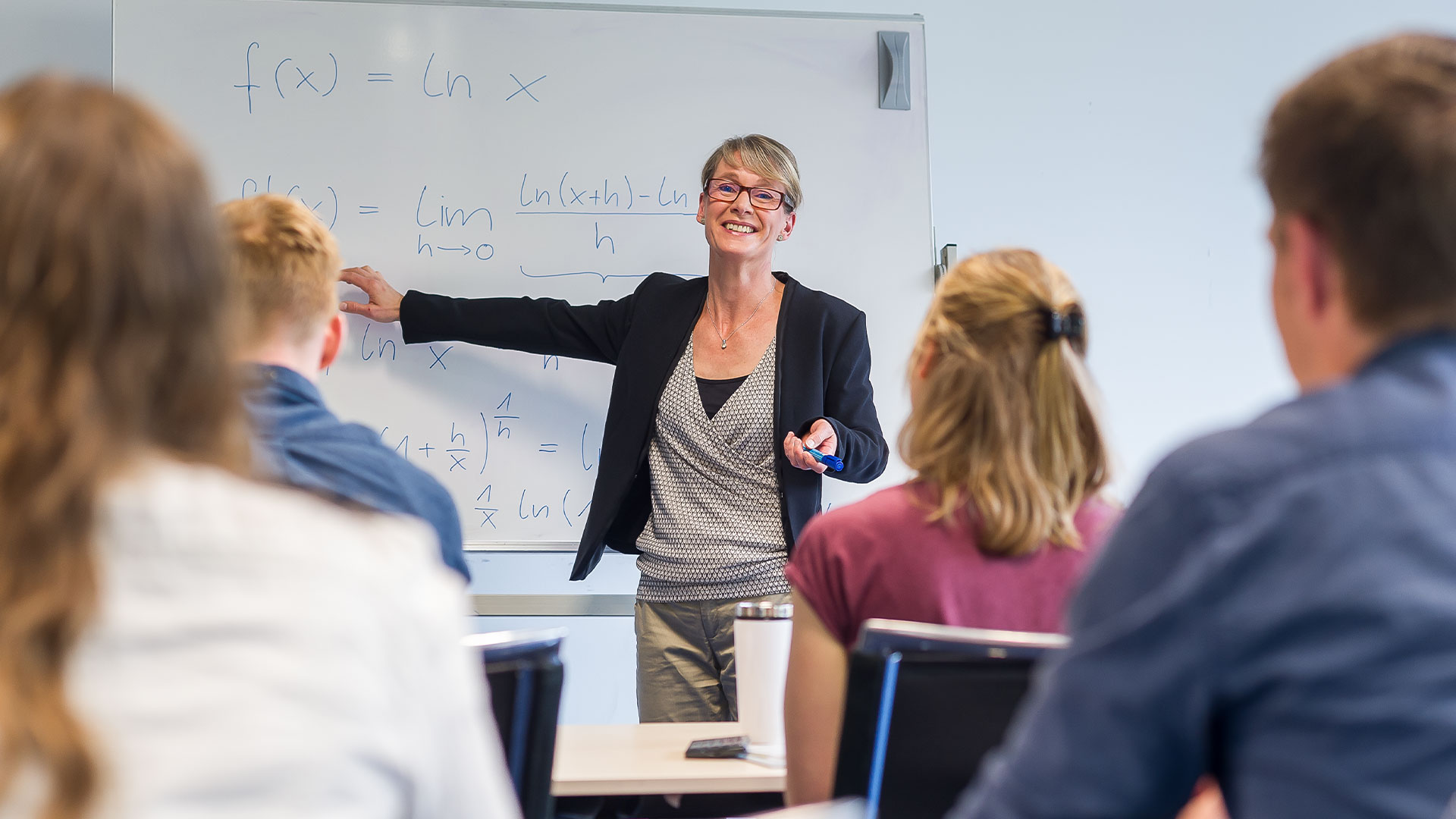 Eine Frau zeigt auf ein Whiteboard, auf das sie etwas geschrieben hat. Sie schaut zu ihren Studenten und erklärt etwas.
