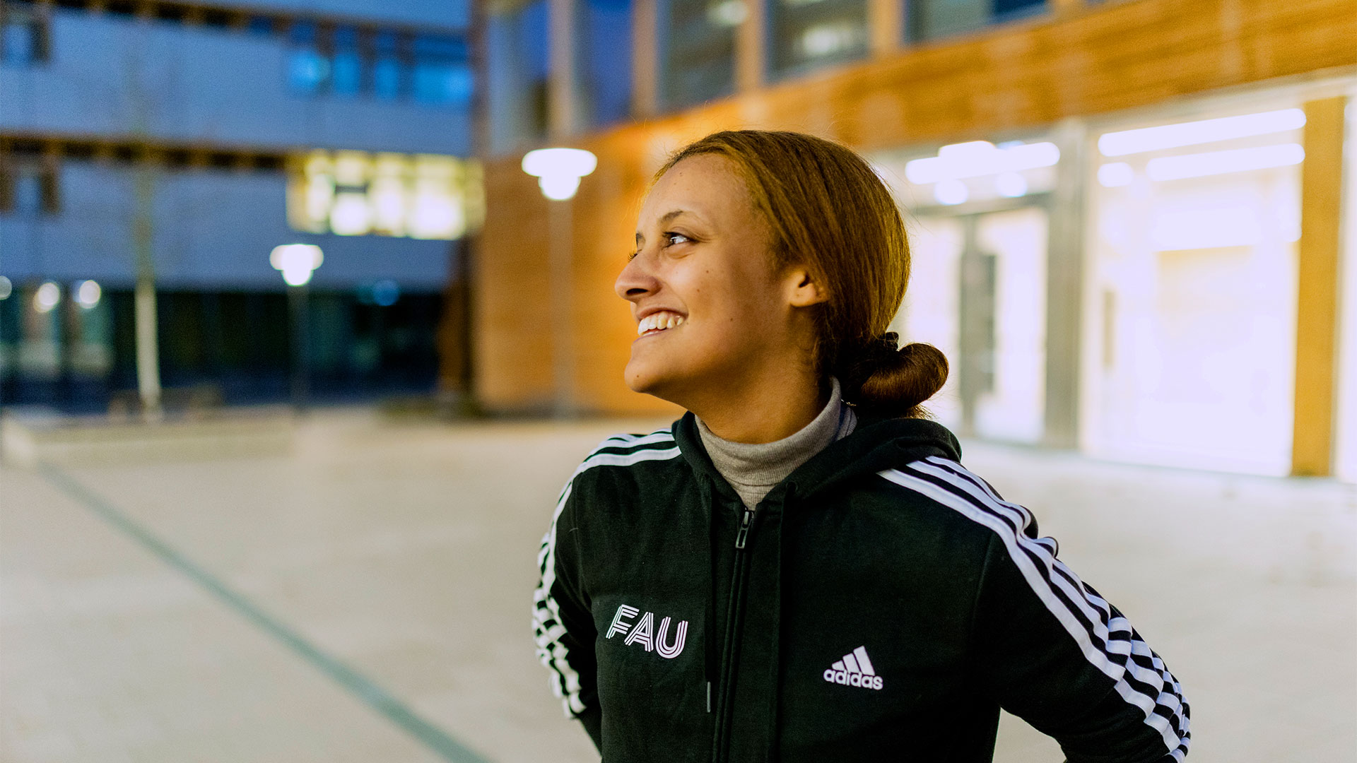 A young woman standing outside at dusk in front of a research building, looking off to the distance and smiling.