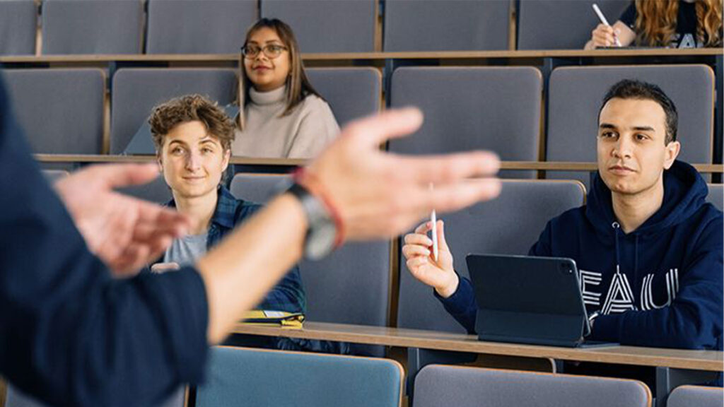 Studentinnen und Studenten sitzen im Hörsaal und diskutieren mit einem Dozenten, von dem man den gestikulierenden Oberarm sieht.