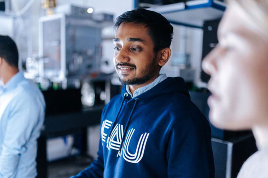 Ein junger Mann mit FAU-Hoodie steht in einem Raum, in dem geforscht wird. Links und rechts von ihm befindet sich jeweils eine weitere Person